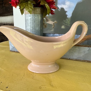 May include: A pale pink ceramic gravy boat with a single handle. The boat is sitting on a yellow wooden surface.