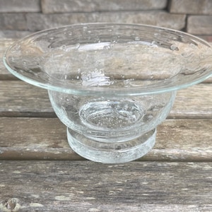 May include: A clear glass bowl with a wide, flared rim and a pedestal base. The bowl has a slightly textured surface with small bubbles throughout. The bowl is sitting on a wooden surface.