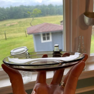 May include: A clear glass saucer with a slight purple tint. The saucer is being held in a hand.