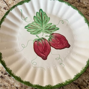 May include: A white ceramic plate with a ruffled edge and a green trim. The plate features a hand-painted design of two red strawberries and a green leaf with delicate green vines.