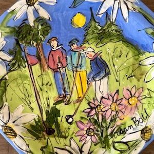 May include: A round ceramic plate featuring a hand-painted scene of three people playing golf on a green course. The artwork includes white and pink daisies, a yellow sun, and trees against a blue sky. The artist signature Susan Boll is visible.