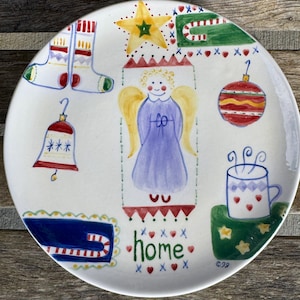 Può includere: Un piatto in ceramica bianca con illustrazioni natalizie dipinte a mano. Il piatto presenta una campana, un angelo, una stella, un ornamento, una tazza e la parola "home". Il piatto ha uno stile rustico e popolare.