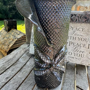 Puede incluir: Jarrón de cerámica negro con una superficie texturizada y un gran lazo. El jarrón tiene una forma única y asimétrica.