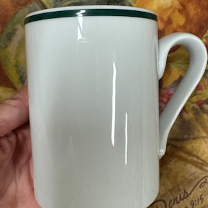 May include: White ceramic mug with a green rim and a handle. The mug is empty and has a glossy finish.