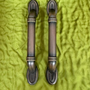 May include: Two matching cabinet pulls with a dark bronze finish. Each handle features a cylindrical center bar with three horizontal ribbed bands and oval-shaped mounting plates at the top and bottom, displaying a concentric ring design.