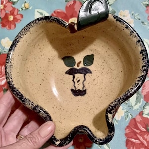 May include: A ceramic apple-shaped bowl with a black rim and a painted apple design in the centre. The bowl has a brown handle shaped like a leaf.