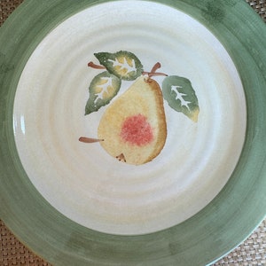 Puede incluir: Un plato de cena de cerámica blanca con un borde verde y un diseño de pera pintado a mano. La pera es amarilla con un rubor rojo y hojas verdes.