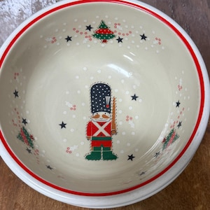 May include: Cream-colored ceramic bowl with a red rim, featuring a festive design. The bowl's interior showcases a toy soldier, Christmas tree, stars, and snowflakes. A charming holiday-themed decorative piece.