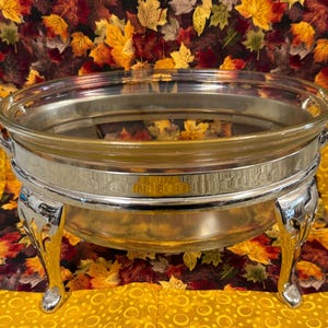 May include: A silver chafing dish with a clear glass insert and ornate handles and legs. The dish sits on a yellow and fall-themed patterned tablecloth. Ideal for serving food at a buffet or party.