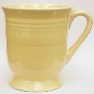 May include: A pale yellow ceramic mug with a curved handle and a pedestal base. The mug has a smooth, glossy finish and horizontal ridges around the middle. The mug is designed for hot beverages.