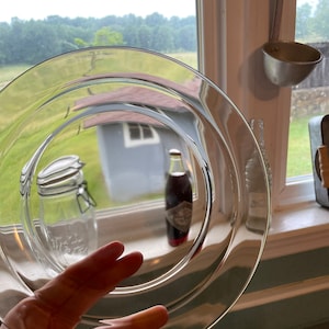 May include: A clear glass plate with a slightly rippled edge. The plate is being held by a hand, and the reflection of a glass jar and a bottle can be seen in the plate.