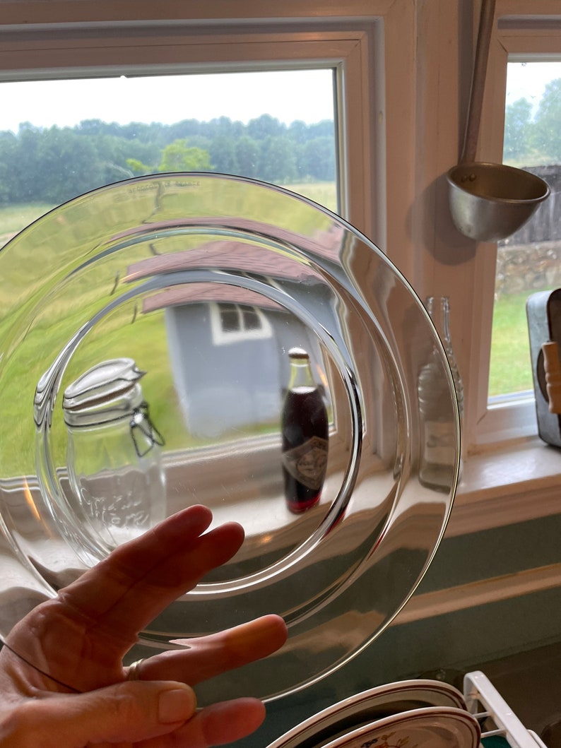 May include: A clear glass plate with a slightly rippled edge. The plate is being held by a hand, and the reflection of a glass jar and a bottle can be seen in the plate.