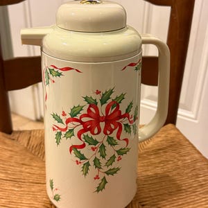 Puede incluir: Termo aislado de color crema con un diseño de lazo rojo y acebo. El termo tiene un asa y un pico vertedor. La tapa también es de color crema con un botón dorado. El termo está sobre una silla de mimbre.