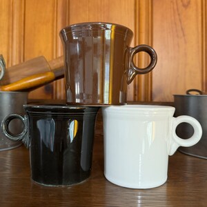 Puede incluir: Tres tazas de cerámica en diferentes colores: marrón, negro y blanco. Las tazas están apiladas una encima de la otra.
