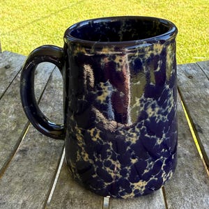 May include: A large, navy blue ceramic mug with a speckled, cream-colored pattern. The mug has a rounded body, a sturdy handle, and a glossy finish. The mug is sitting on a weathered wooden surface.