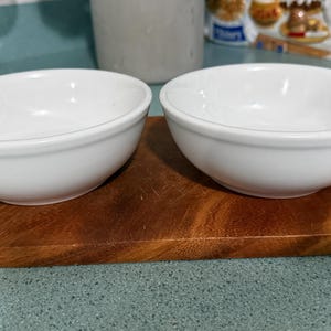 May include: Two white ceramic bowls sit side-by-side on a wooden cutting board. The bowls are round with a simple, classic design. The wooden board has a rich, brown color, and the background is a light blue countertop.