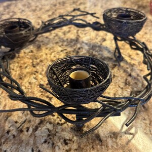 May include: A black metal candle holder shaped like a wreath with three bird's nest candle holders. The candle holder is made of twisted metal branches.