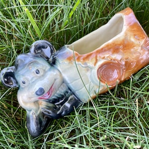 Puede incluir: Una maceta de cerámica con forma de tronco de árbol con una cara de oso de dibujos animados en el lateral. La maceta está esmaltada en marrón, blanco y azul.