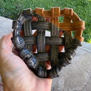 May include: A ceramic coaster with a woven design. The coaster features a mix of colors, including shades of gray, brown, and orange. The coaster is held in a hand, with a green grassy background.