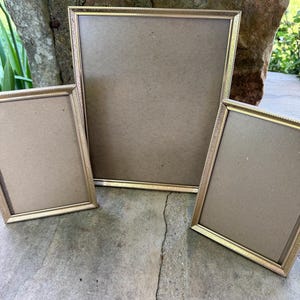 May include: A set of three rectangular picture frames with thin, metallic gold-toned borders featuring a textured, beaded edge detail. The frames are empty, displaying plain brown backing boards, and are arranged in a cluster with one large frame.