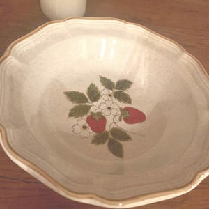 May include: A white ceramic bowl with a brown rim and a strawberry design. The bowl has a scalloped edge and is decorated with green leaves, white flowers, and red strawberries.