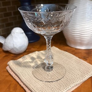 May include: A clear glass coupe with a wide, shallow bowl featuring an etched leaf and floral pattern. The stem has a textured, diamond-cut design leading down to a smooth, circular base. The glass is transparent with intricate detailing.