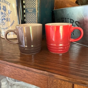 Puede incluir: Dos tazas de cerámica, una marrón y otra roja, con un borde y un asa blancos. Las tazas están sobre una superficie de madera.