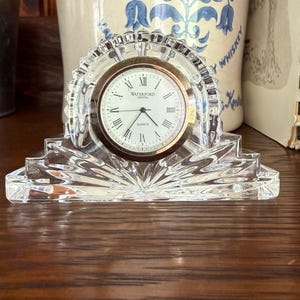 Puede incluir: Un reloj de cuarzo de cristal transparente con esfera dorada y números romanos. El reloj está en un marco de cristal decorativo en forma de abanico. La esfera del reloj dice "Waterford Crystal Quartz".