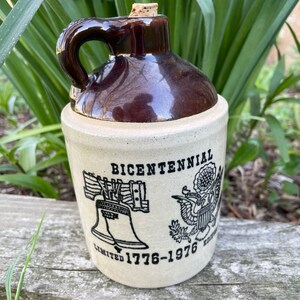 Könnte beinhalten: Eine braune und beigefarbene Keramikkanne mit einem Korkstopfen. Die Kanne hat ein schwarzes Design mit den Worten "Bicentennial Limited 1776-1976 Edition" und Bildern der Freiheitsglocke und des amerikanischen Adlers.