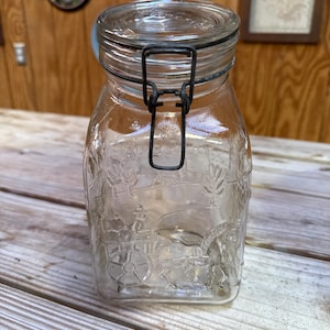 Puede incluir: Un tarro de vidrio transparente con cierre de metal. El tarro tiene forma cuadrada y presenta un diseño en relieve de un carruaje tirado por caballos y árboles. El tarro está sobre una superficie de madera.