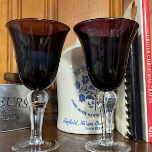Puede incluir: Dos copas de vino antiguas con cuencos burdeos oscuros y tallos transparentes y ornamentados. Las copas están sobre una superficie de madera, con una jarra de cerámica y un libro titulado "Glorious Christmas Crafts" en el fondo.