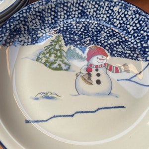May include: A white ceramic bowl with a blue and white speckled glaze. The bowl features a winter scene with a snowman wearing a red hat and scarf, holding a red mitten and ice skates. The bowl is perfect for serving soup, salad, or dessert.
