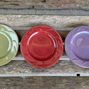 May include: Three ceramic plates in a row, each with a different colour and design. The plates are green, red, and purple. Each plate has a leaf pattern around the rim.