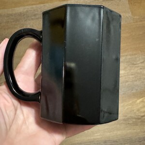 May include: A black ceramic mug with a hexagonal shape. The mug has a handle and is made of a smooth, glossy material.