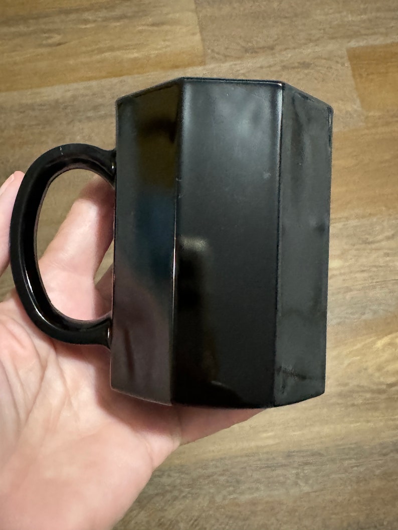 May include: A black ceramic mug with a hexagonal shape. The mug has a handle and is made of a smooth, glossy material.