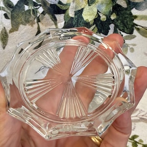 Op de afbeelding: Heldere, achthoekige glazen asbak met een sterrenpatroon in het midden. De asbak heeft een facetrand en wordt in een hand gehouden. De achtergrond is een bloemenpatroon.