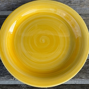 Puede incluir: Un plato de cerámica amarillo brillante con un diseño en espiral. El plato tiene un borde ancho y un centro ligeramente cóncavo. El color es un amarillo vibrante y soleado, con sutiles variaciones de tono. Adecuado para servir alimentos.