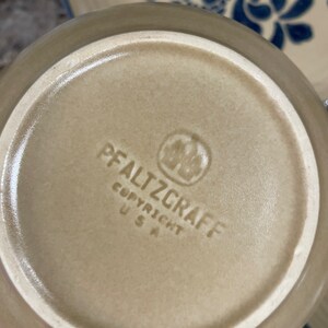 May include: The bottom of a tan ceramic plate with a blue and white floral design. The plate has the words "Pfaltzgraff Copyright USA" printed on the bottom.