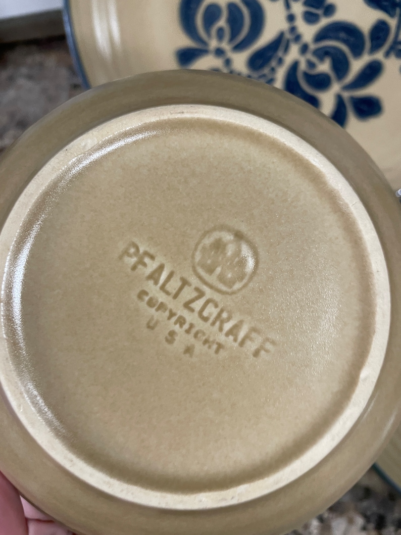 May include: The bottom of a tan ceramic plate with a blue and white floral design. The plate has the words "Pfaltzgraff Copyright USA" printed on the bottom.