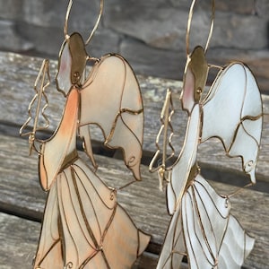 Peut inclure: Deux figurines d'anges debout composées de panneaux translucides blanc cassé et pêche, encadrés par un fil métallique doré. Chaque ange possède une auréole circulaire, des ailes déployées et une longue robe fluide, tenant un bâton surmonté d'une étoile.