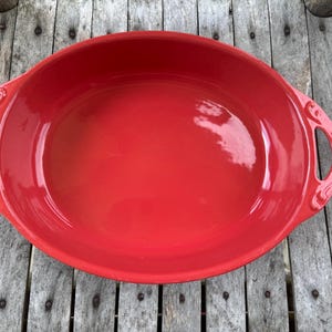 Puede incluir: Un plato para hornear de cerámica ovalado de color rojo con dos asas. El plato está vacío y tiene un acabado brillante.