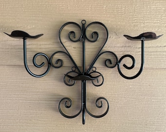 May include: Black metal wall sconce with a heart-shaped design and three candle holders. The sconce features decorative scrollwork and a matte finish, ideal for adding a touch of elegance to any room. The candle holders are shaped like small dishes.