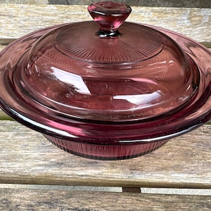 Può includere: Una pirofila vintage in vetro viola scuro con coperchio abbinato. Il coperchio presenta un pomello centrale e un design a coste che si irradia verso l'esterno. La pirofila e il coperchio sono in vetro trasparente. La pirofila è su una superficie di legno.