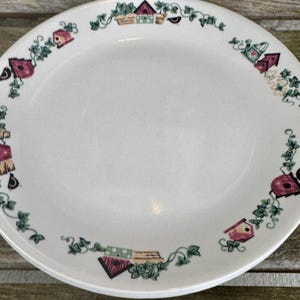 May include: White ceramic plate with a decorative border featuring birdhouses, baskets, and vines in red, green, and brown. The plate has a slightly textured surface and a simple design, suitable for everyday use.