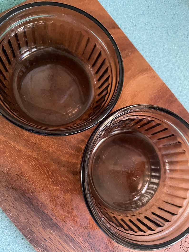 May include: Two brown glass ramekins with fluted edges. The ramekins are empty and sitting on a wooden surface.