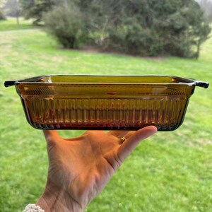 May include: A rectangular amber-coloured glass baking dish featuring vertical ribbed detailing along the exterior walls and two small integrated handles on opposite sides. The dish is translucent and has a smooth, flat rim around the top edge.