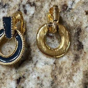 May include: A pair of gold tone clip-on earrings with black enamel accents. The earrings are round and have a ring design.