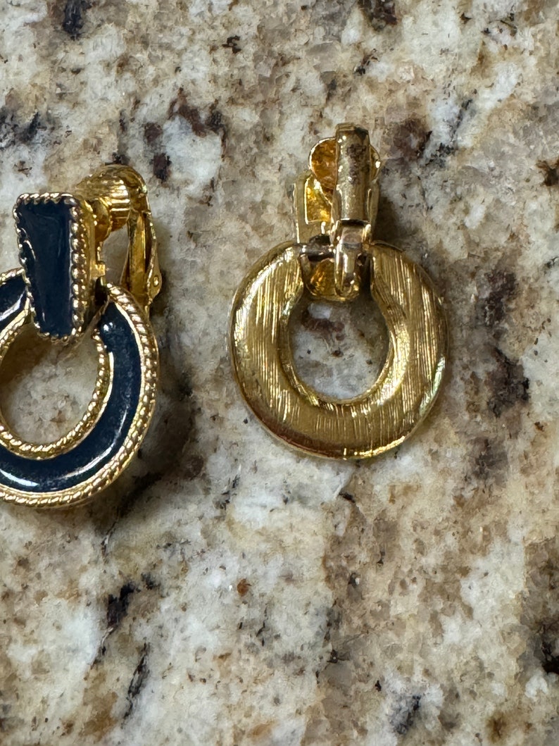 May include: A pair of gold tone clip-on earrings with black enamel accents. The earrings are round and have a ring design.
