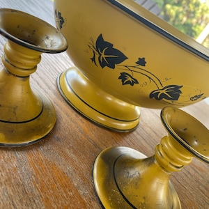 May include: Three yellow metal candle holders with black trim. The candle holders have a simple design with a wide base and a narrow top. The candle holders are on a wooden surface.