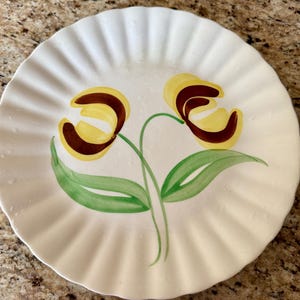May include: White ceramic plate with a ruffled edge, hand-painted with two yellow and brown tulip flowers and green leaves. The plate has a vintage aesthetic.